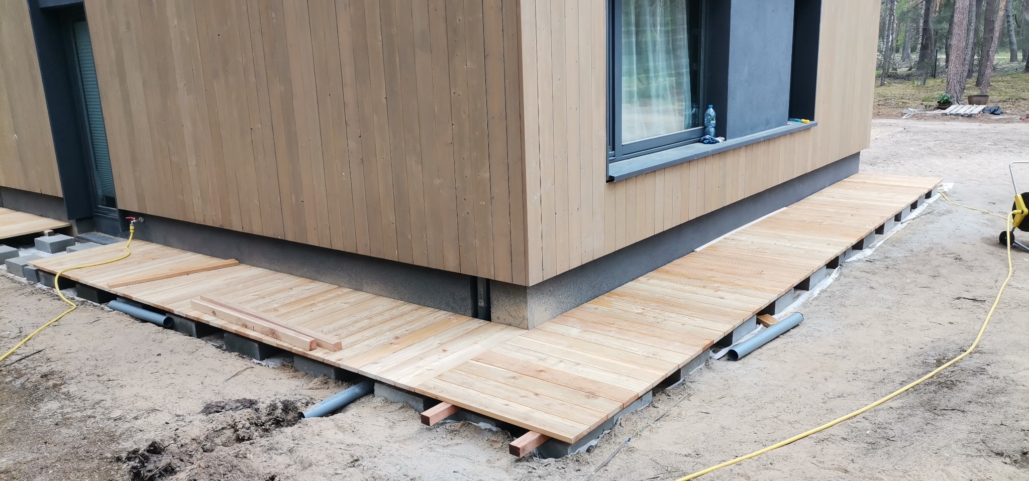 Budowa tarasu drewnianego przy nowoczesnym domu, widoczne elementy konstrukcyjne, rury drenażowe i żółty wąż ogrodowy.