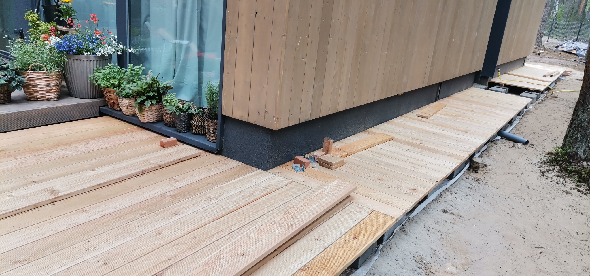 Montaż drewnianego tarasu przy nowoczesnym domu z kwiatami w doniczkach, widoczne elementy konstrukcyjne i narzędzia.