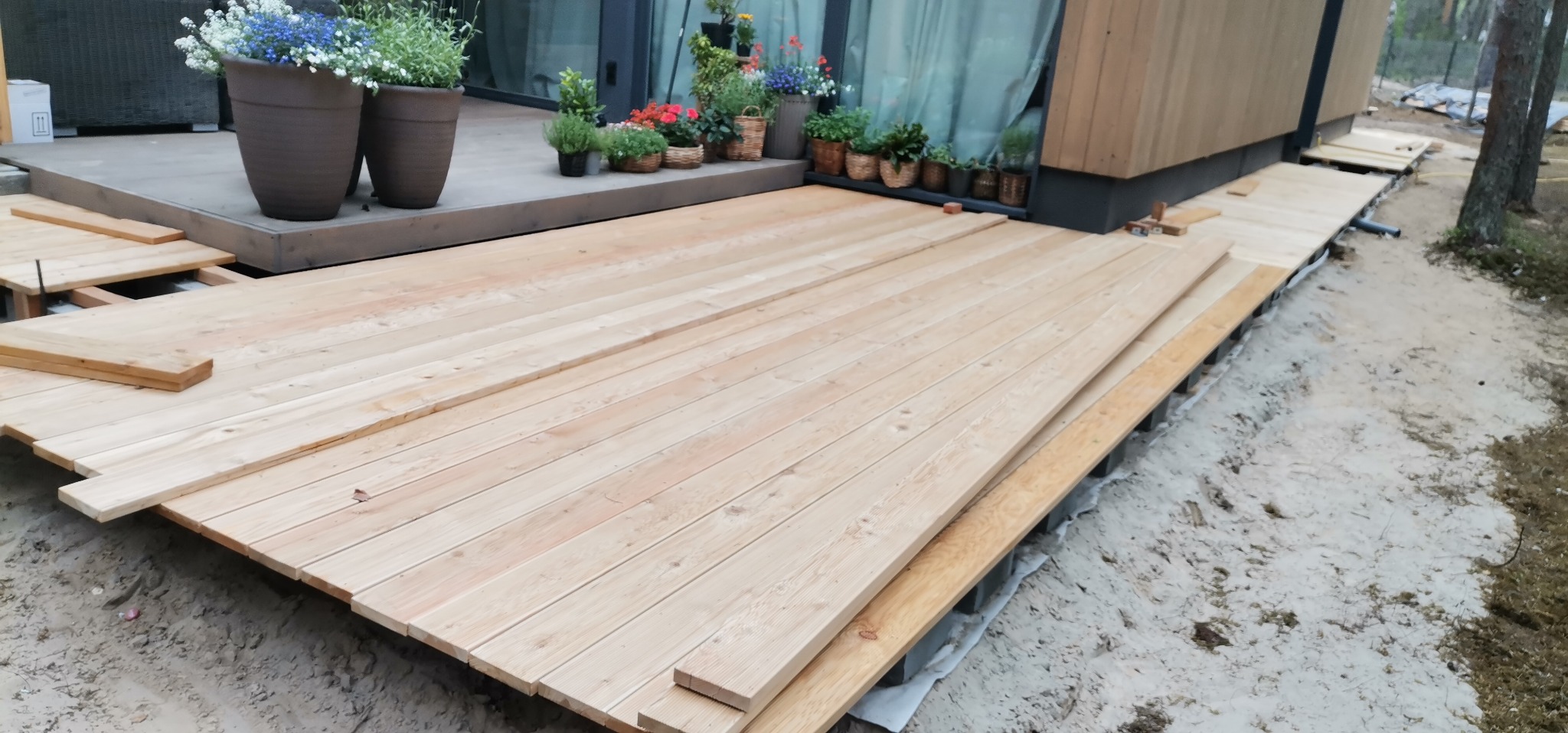 Montaż tarasu drewnianego z jasnych desek na podłożu z piasku, widoczne donice z kwiatami na istniejącej części tarasu.