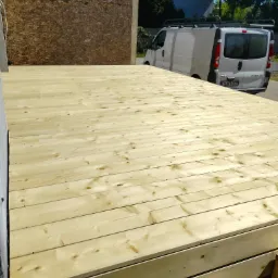 Nowy taras z jasnego drewna, widok z góry na deski, z tyłu zaparkowany biały bus.