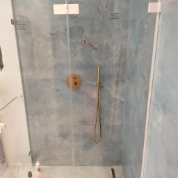 Artbud Kubiak - Kabina prysznicowa z deszczownicą i baterią w kolorze złotym, wykończona płytkami imitującymi marmur z delikatnym niebieskim odcieniem, widoczne akcesoria kosmetyczne na podłodze.