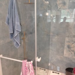 Artbud Kubiak - Nowoczesna kabina prysznicowa z deszczownicą i szklanymi drzwiami, wykończona marmurowymi płytkami w odcieniach szarości i błękitu, z wiszącym jasnoniebieskim ręcznikiem i ubraniami w paski...