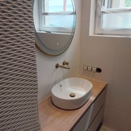 Artbud Kubiak - Umywalka nablatowa owalna z baterią w kolorze szczotkowanego złota, zamontowana na drewnopodobnym blacie z frezowanymi frontami szafki, okrągłe lustro z podświetleniem LED, białe płytki ścienne...