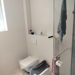 Artbud Kubiak - Biała łazienka z wiszącą szarą kabiną prysznicową i toaletą; na półce kosmetyki i akcesoria, widoczny skrobak do szyb.