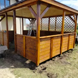 Nowo wybudowana, drewniana altana z kratką, częściowo osłonięta drewnianymi panelami, z drabiną wewnątrz, na trawniku przed domem w słoneczny dzień.