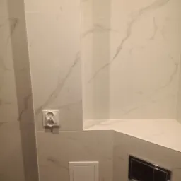 Wykończona ściana w łazience z białych płytek imitujących marmur, widoczne gniazdko elektryczne, przycisk spłuczki i wnęka.