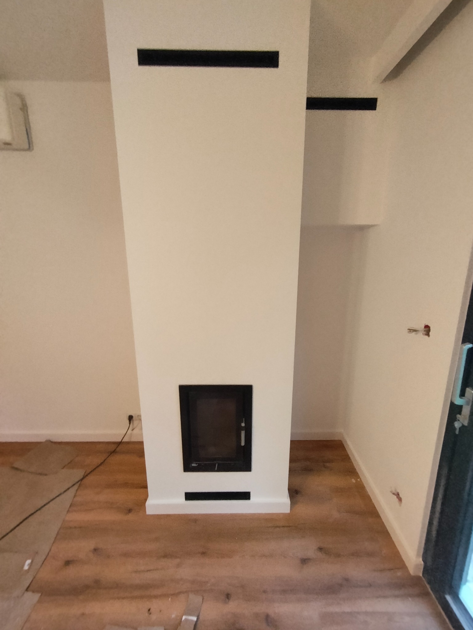 Biały kominek z czarnymi kratkami wentylacyjnymi w nowo wyremontowanym wnętrzu z drewnianą podłogą, widoczne niedokończone prace i zabezpieczenia podłogi.