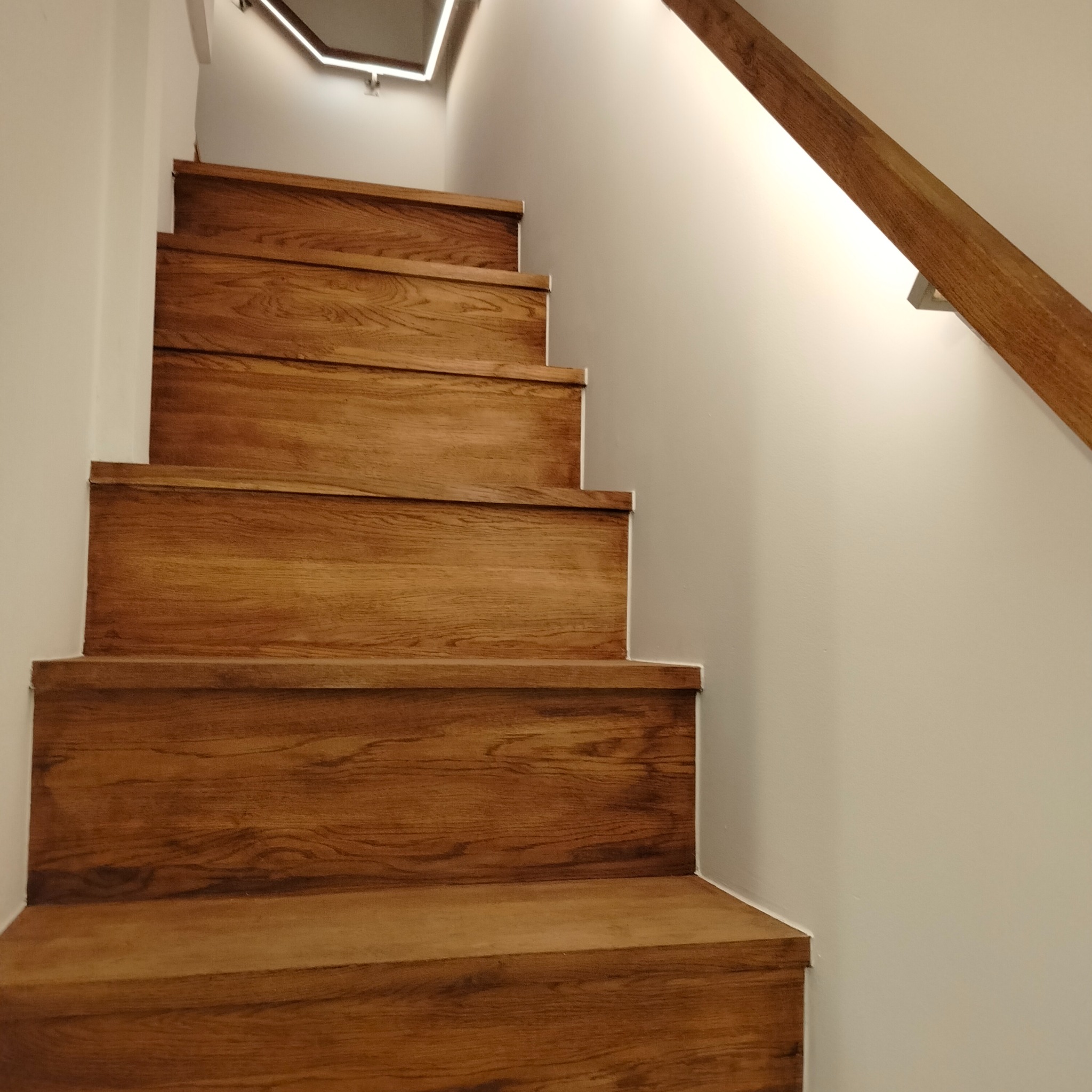 Schody z drewnianymi stopniami, oświetlenie LED pod stopniami i na suficie, drewniana poręcz przy ścianie.
