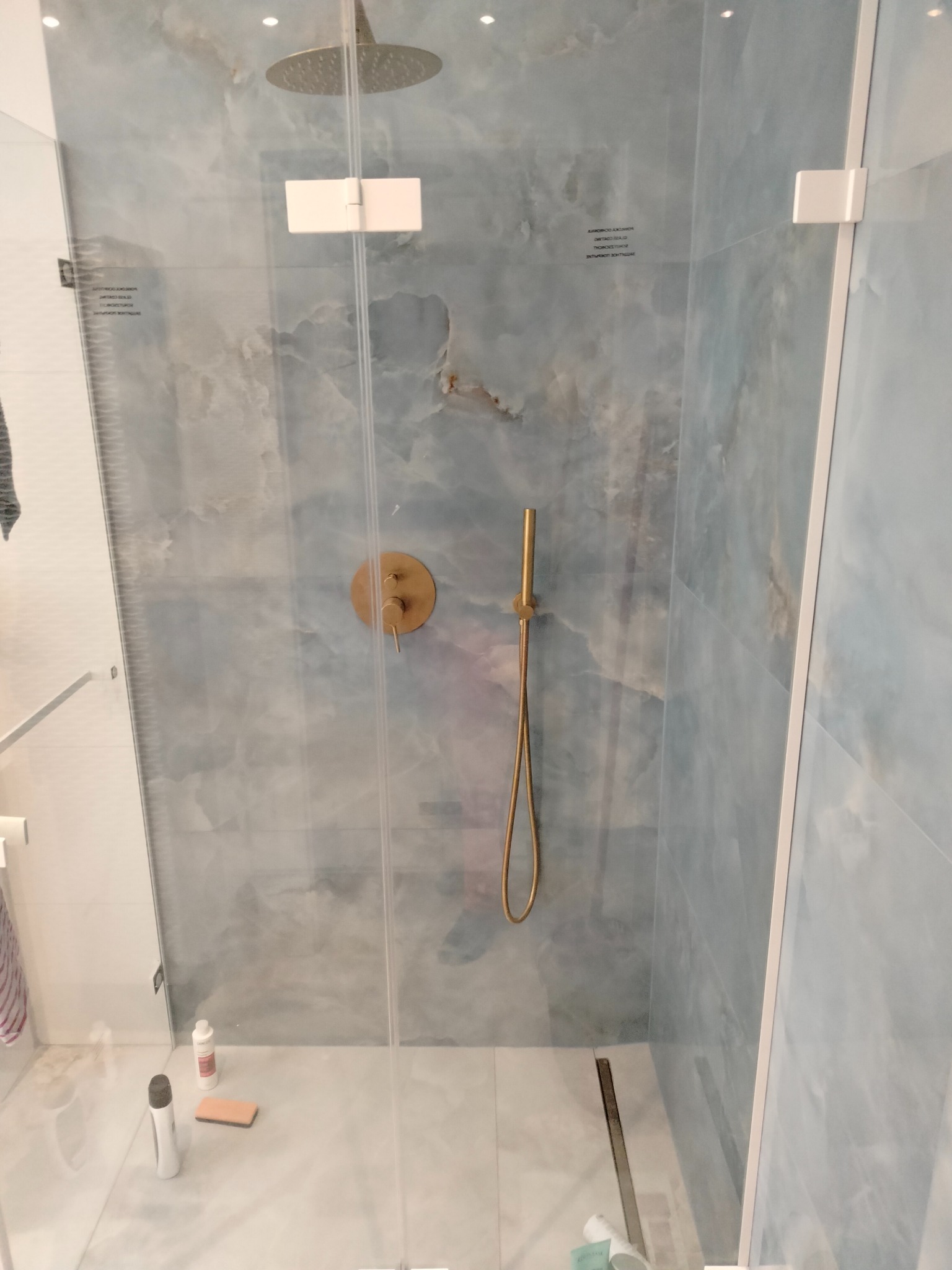 Kabina prysznicowa z deszczownicą i baterią w kolorze złotym, wykończona płytkami imitującymi marmur z delikatnym niebieskim odcieniem, widoczne akcesoria kosmetyczne na podłodze.