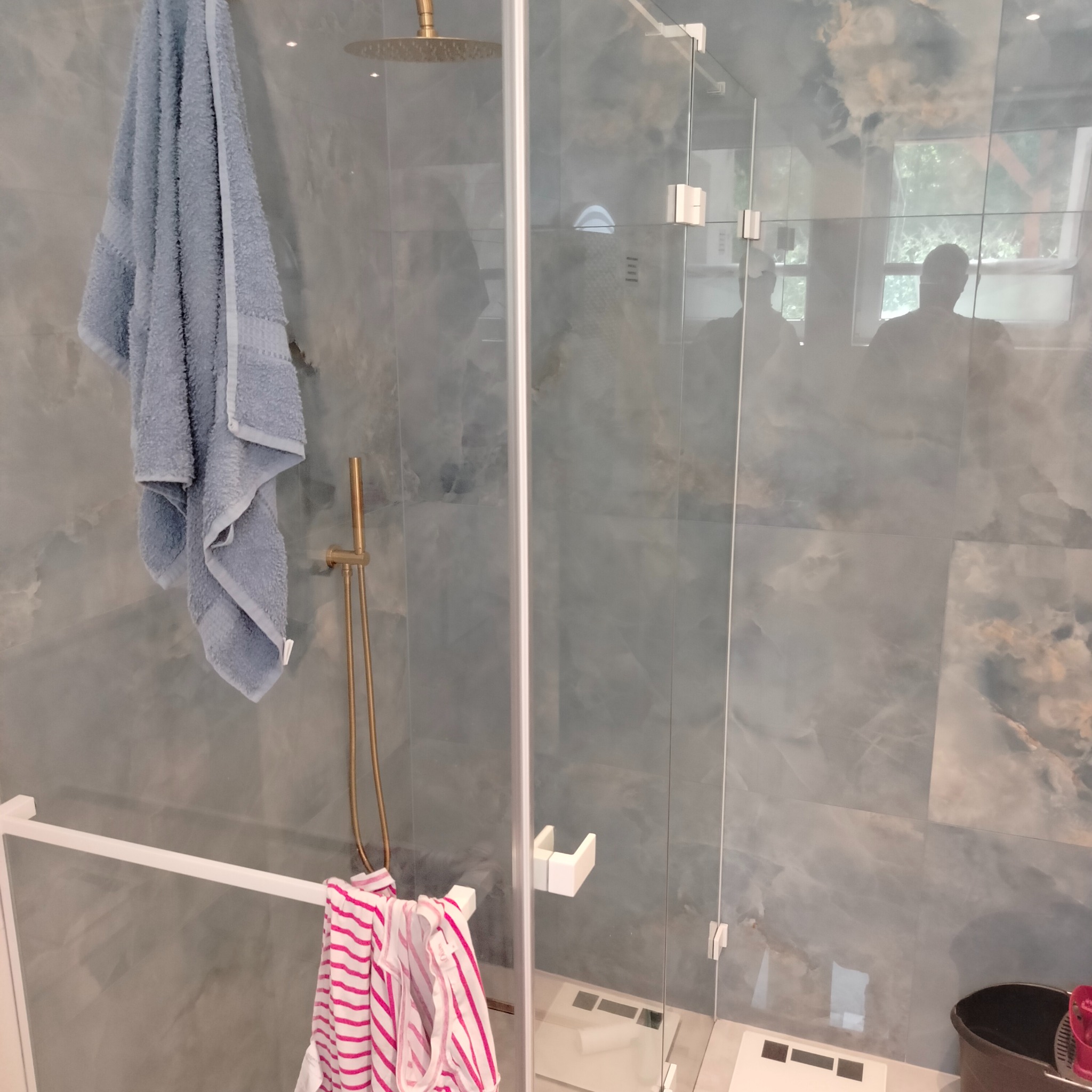 Nowoczesna kabina prysznicowa z deszczownicą i szklanymi drzwiami, wykończona marmurowymi płytkami w odcieniach szarości i błękitu, z wiszącym jasnoniebieskim ręcznikiem i ubraniami w paski...