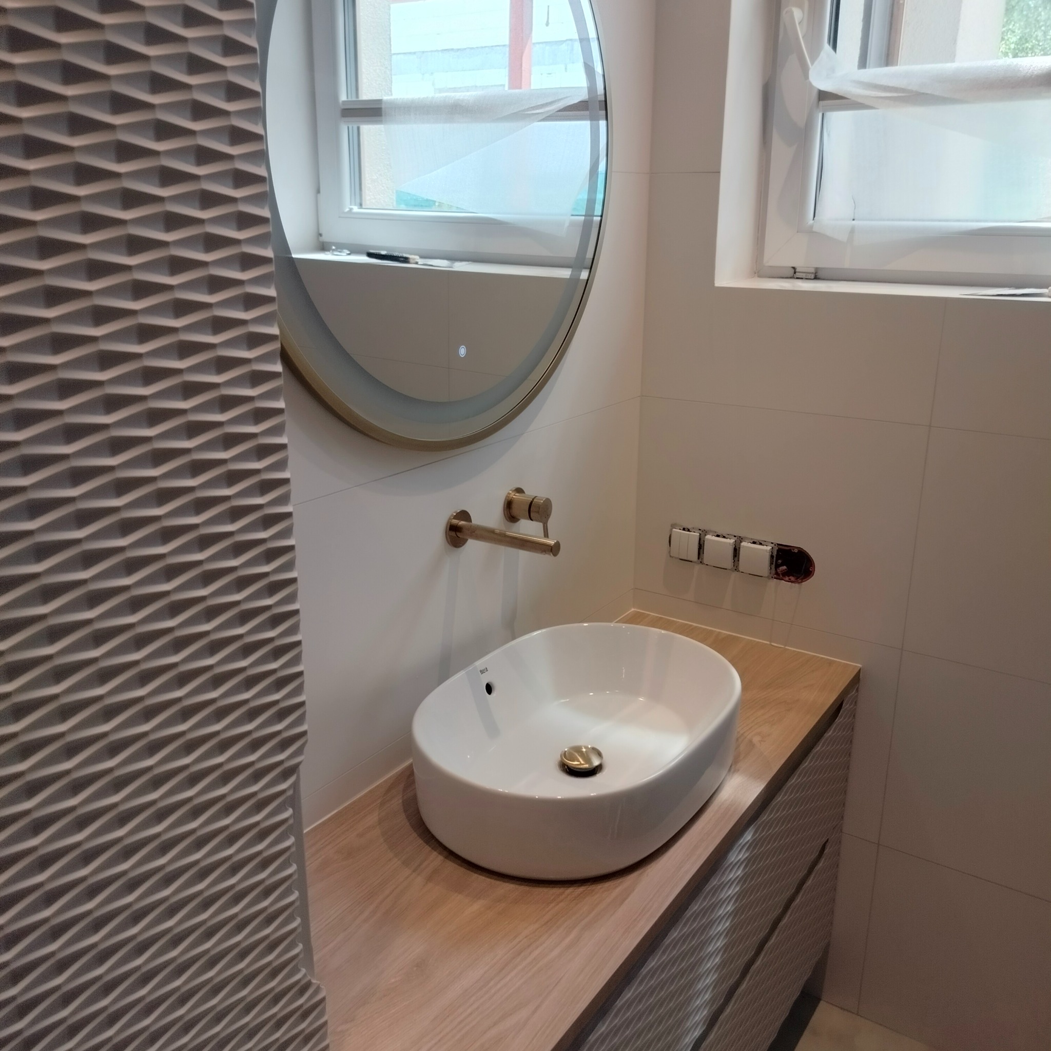 Umywalka nablatowa owalna z baterią w kolorze szczotkowanego złota, zamontowana na drewnopodobnym blacie z frezowanymi frontami szafki, okrągłe lustro z podświetleniem LED, białe płytki ścienne...