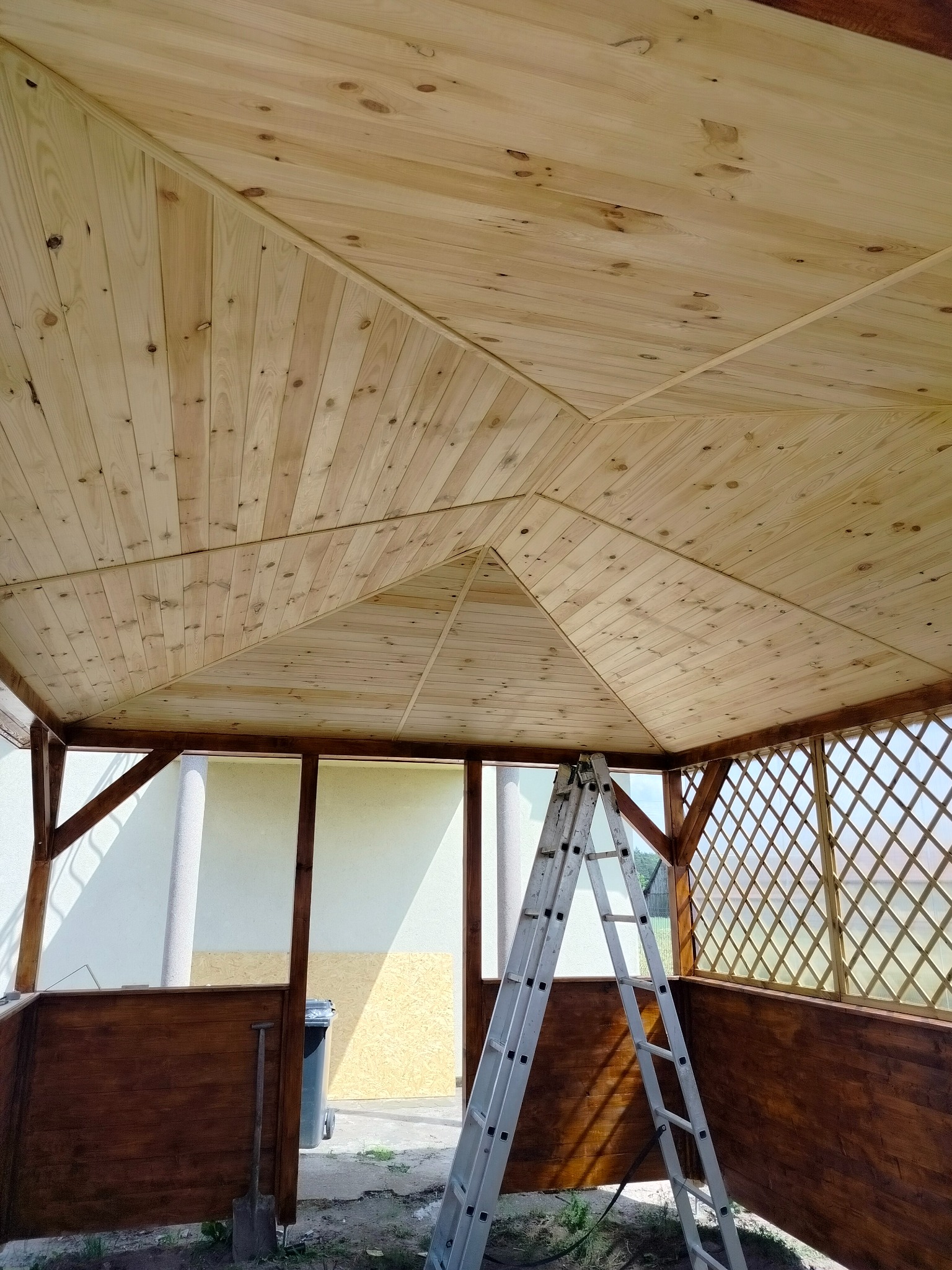 Wnętrze altany w trakcie budowy, widoczny drewniany sufit z desek, pomalowane na brązowo ściany z kratownicą i drabina oparta o ścianę.