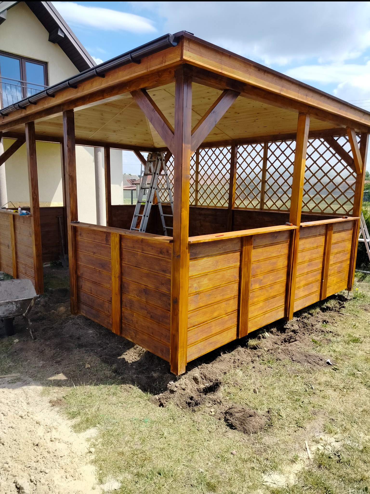 Nowo wybudowana, drewniana altana z kratką, częściowo osłonięta drewnianymi panelami, z drabiną wewnątrz, na trawniku przed domem w słoneczny dzień.