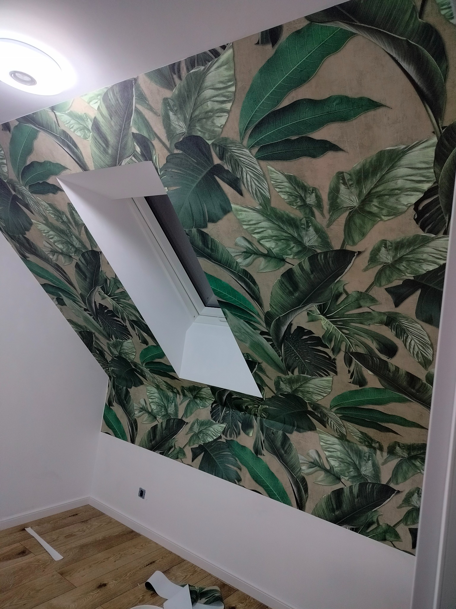 Pokój na poddaszu z oknem dachowym, ściana pokryta tapetą w motywy liści tropikalnych, jasna podłoga z paneli, fragmenty odciętej tapety na podłodze.
