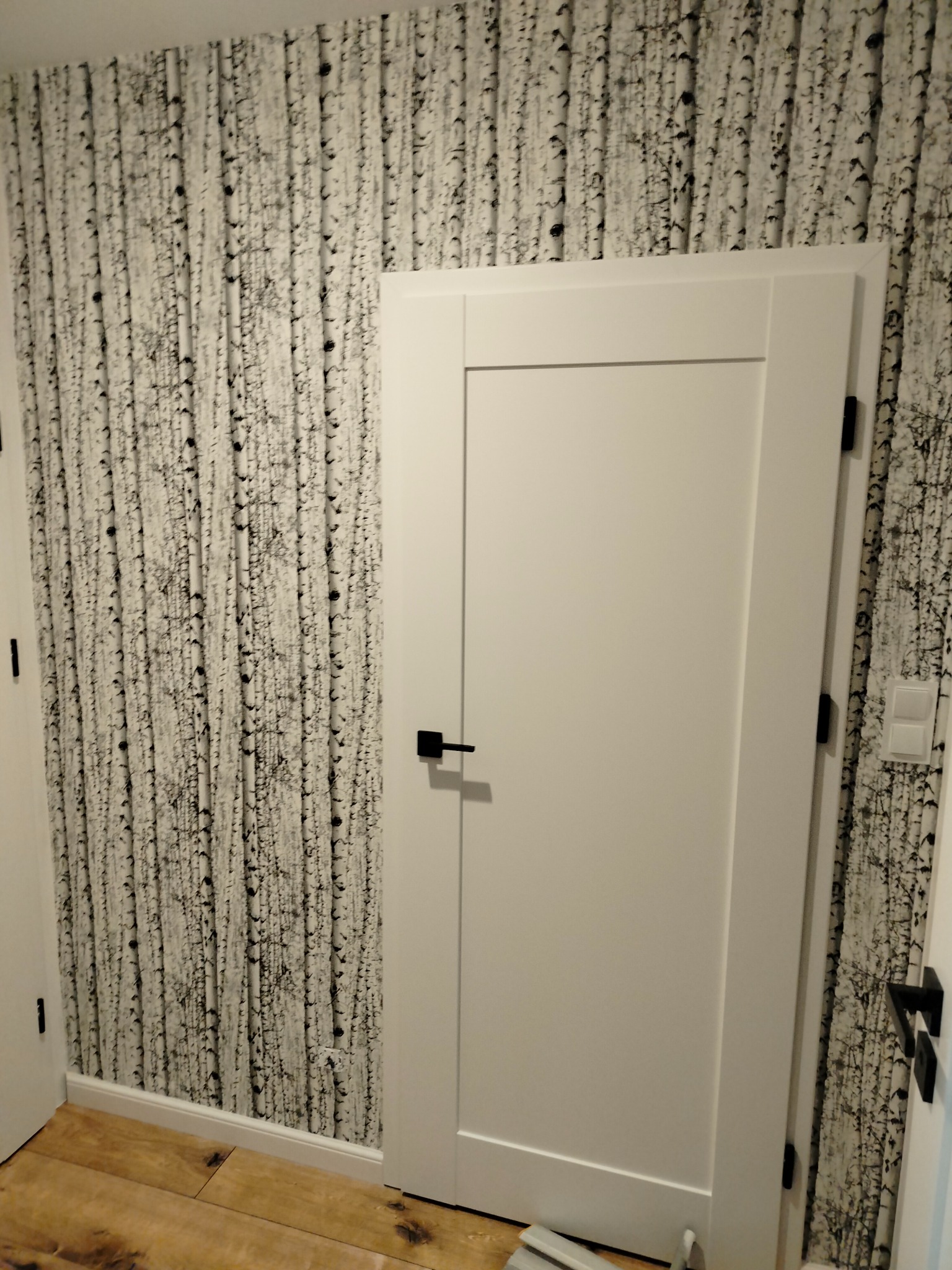 Białe drzwi wewnętrzne z czarną klamką na tle ściany pokrytej tapetą imitującą brzozowy las, widoczne fragmenty drewnianej podłogi.