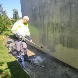 Pracownik w odblaskowej kurtce czyści elewację budynku za pomocą myjki ciśnieniowej Kärcher, widoczne ślady czyszczenia na ścianie i mokra kostka brukowa.