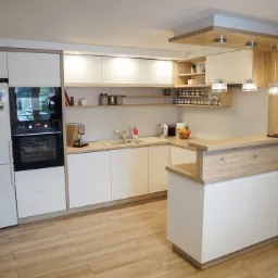 Jasna kuchnia z białymi frontami i drewnianymi akcentami, widoczne półki z przyprawami, ekspres do kawy i lodówka z magnesami, oświetlenie punktowe i drewniana podłoga.