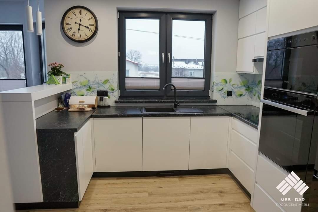 Nowoczesna kuchnia w kształcie litery U z białymi frontami szafek i blatem w kolorze czarnego marmuru, zlew pod oknem, pomiędzy szafkami nad blatem panel z motywem limonki, na ścianie zegar...