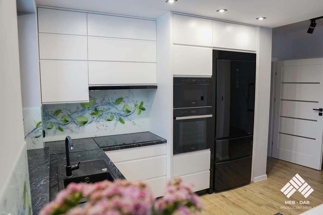 Nowoczesna biała zabudowa kuchenna z czarnym blatem i grafiką z motywem limonki na szklanym panelu ściennym, zintegrowana z czarnym piekarnikiem i lodówką, widoczny fragment zlewu i kwiatów.