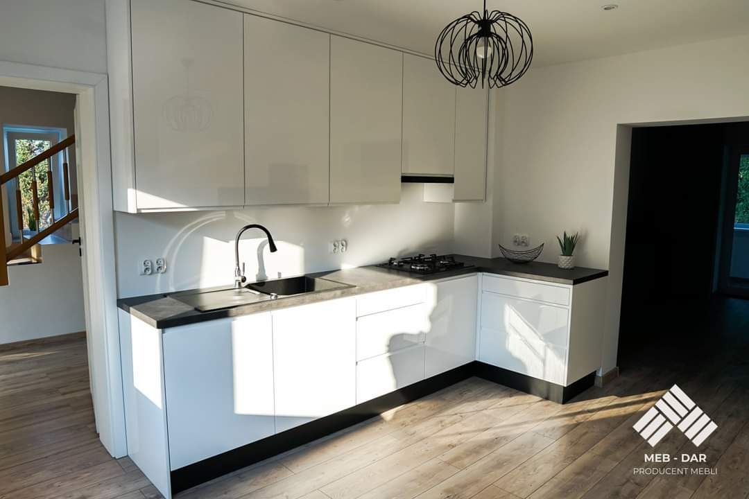 Nowoczesna, biała kuchnia z czarnym blatem i minimalistyczną lampą wiszącą. Widoczny zlew, płyta gazowa i drewniana podłoga. W tle schody z drewnianymi barierkami i logo producenta mebli w prawym...