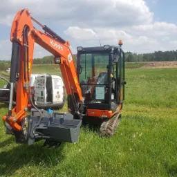 Pomarańczowa mini koparka Kubota z łyżką chwytakową, stojąca na trawiastym terenie, na tle biały samochód dostawczy z otwartymi drzwiami i las w oddali, słoneczny dzień.