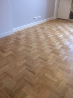 Pusty pokój z nowo wycyklinowaną i polakierowaną parkietową podłogą ułożoną w jodełkę, świeżo pomalowane ściany w odcieniach szarości z białymi listwami przypodłogowymi.