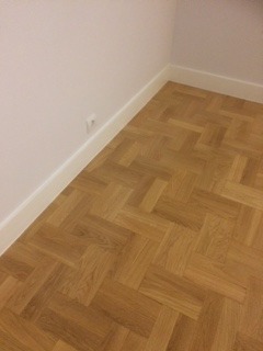 Parkiet ułożony w jodełkę w rogu pomieszczenia, biała listwa przypodłogowa, gładka biała ściana z gniazdkiem elektrycznym.