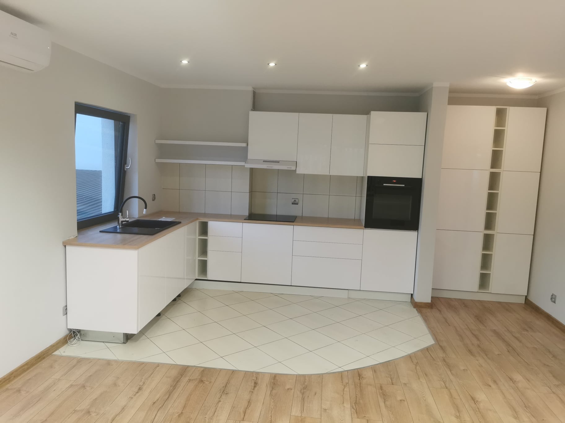 Jasna, nowoczesna kuchnia z białymi frontami szafek, drewnianym blatem i czarnym zlewozmywakiem, na podłodze jasne płytki przechodzące w drewniany parkiet.