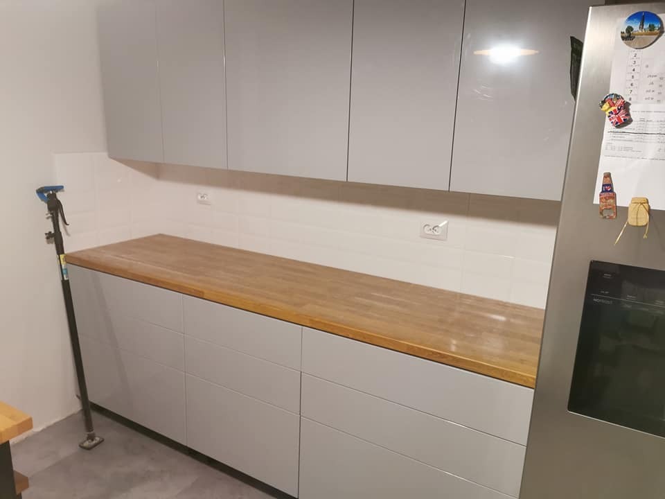 Szare szafki kuchenne z drewnianym blatem, widoczny fragment lodówki z magnesami i rozpórka do montażu, białe płytki na ścianie.