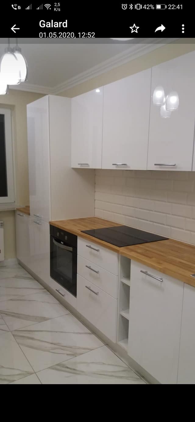 Biała kuchnia na wymiar z drewnianymi blatami i płytą indukcyjną, widok na fronty szafek i piekarnik, płytki w stylu cegiełek na ścianie.