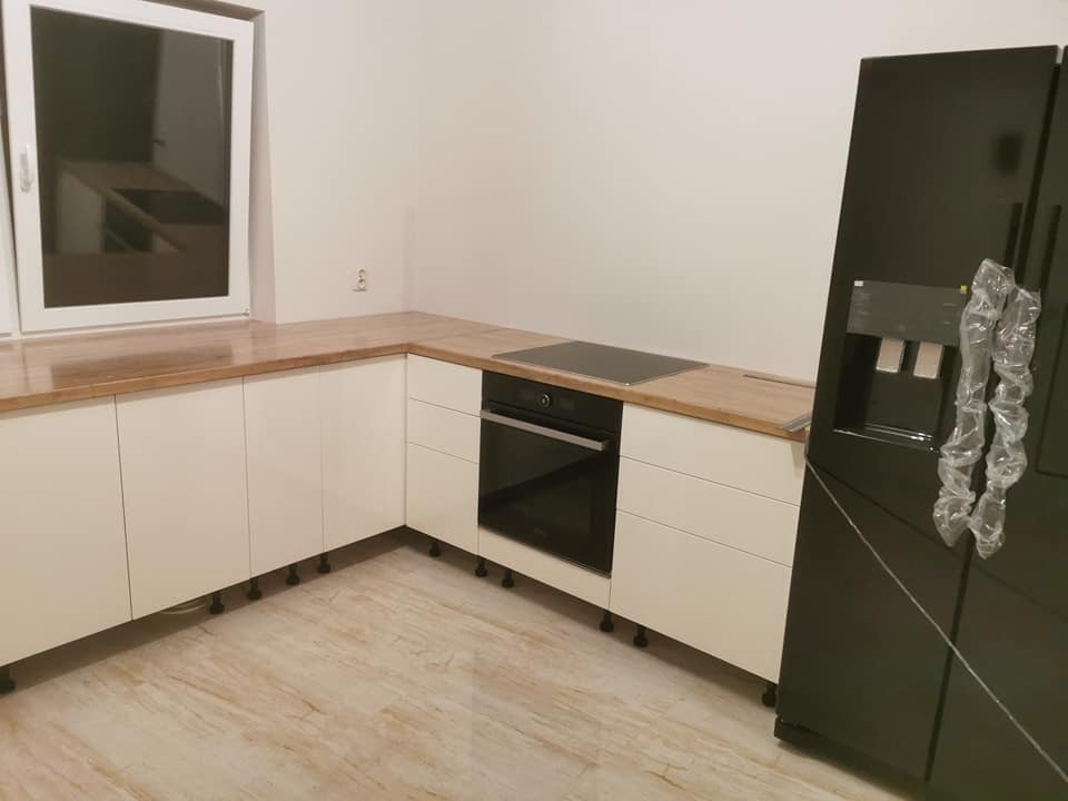 Nowoczesna kuchnia z białymi frontami szafek i drewnianym blatem, czarny piekarnik i lodówka, widok na okno z odbiciem zabudowy kuchennej.