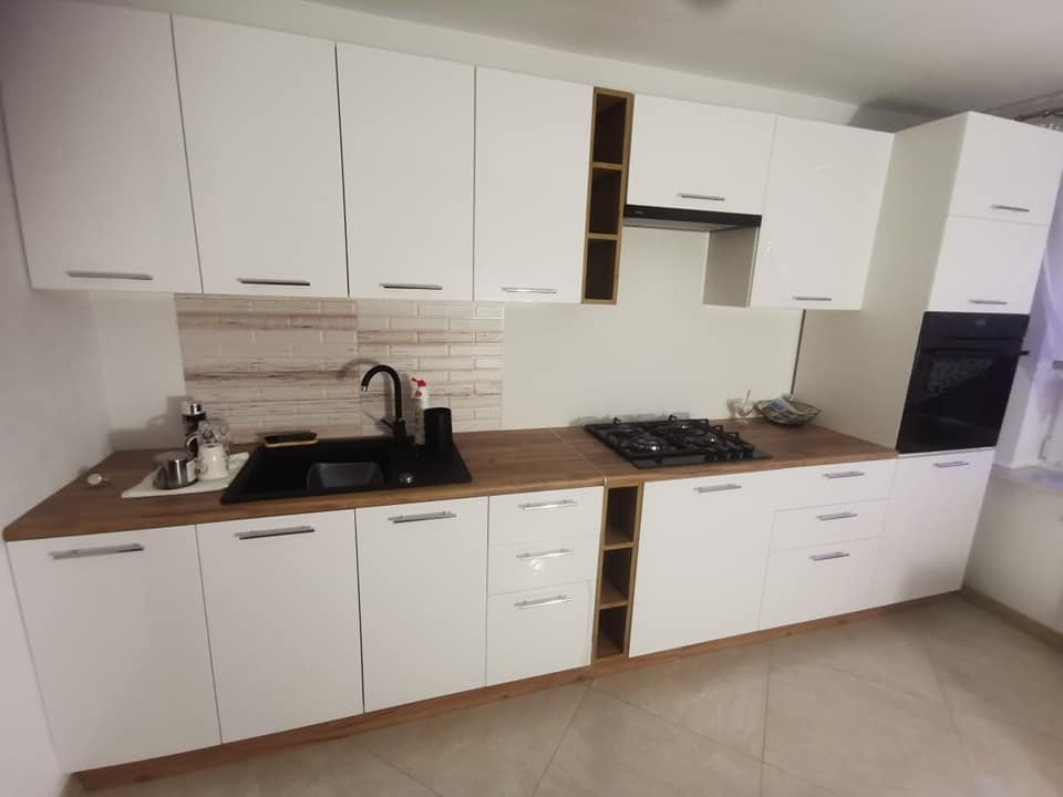 Nowoczesna kuchnia z białymi frontami i drewnianym blatem, czarny zlew i bateria, płyta gazowa, piekarnik w zabudowie, płytki imitujące cegłę na ścianie.