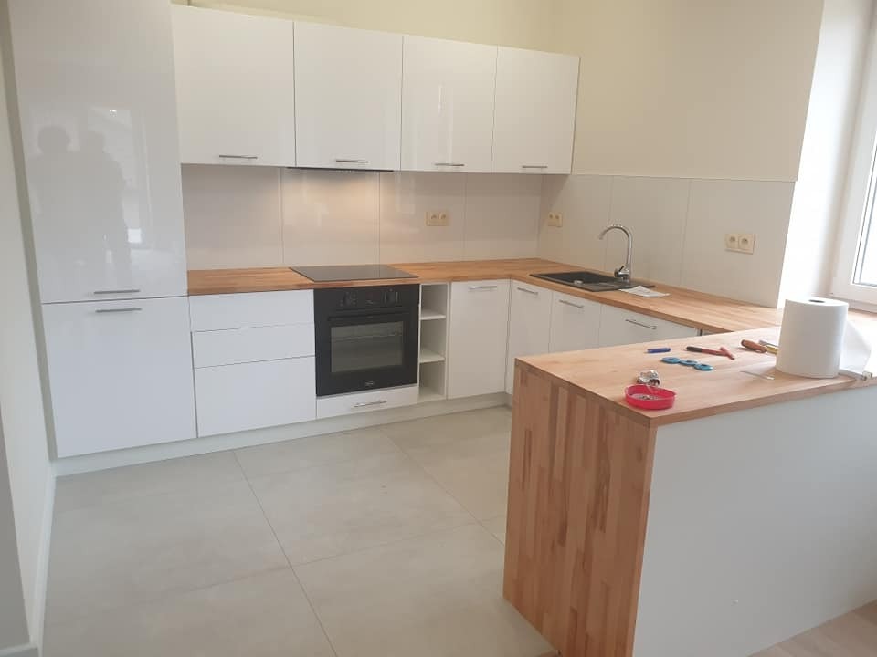 Nowoczesna, jasna kuchnia z białymi, lakierowanymi frontami i drewnianymi blatami; widoczna wyspa kuchenna z akcesoriami montażowymi.