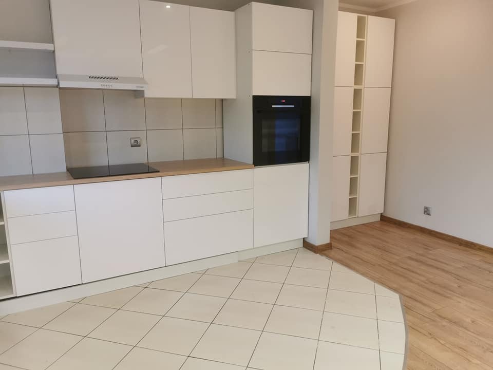 Nowoczesna, jasna kuchnia z białymi, lakierowanymi szafkami, drewnianym blatem i wbudowanym piekarnikiem. Na podłodze kremowe płytki, przechodzące w drewnopodobne panele w sąsiednim pomieszczeniu.