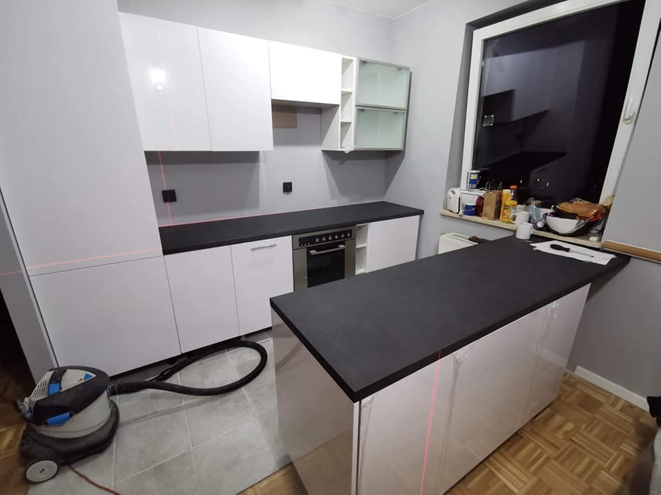 Montaż białej, nowoczesnej kuchni z czarnym blatem, widoczny laserowy poziom i odkurzacz przemysłowy.