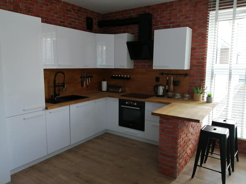 Biała, nowoczesna kuchnia z drewnianym blatem, ceglaną ścianą i barkiem z hokerami, oświetlona naturalnym światłem z okna z żaluzjami.