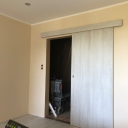 Robert Nawara - Montaż drzwi przesuwnych z szarobiałego drewna w jasnym pokoju, widoczny laser krzyżowy, skrzynki narzędziowe i opakowanie uchwytu ściennego TV na stołku.