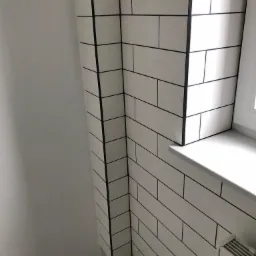 Ściana wyłożona białymi, prostokątnymi płytkami z czarną fugą, narożnik wykończony czarną listwą, widoczny fragment białego parapetu i grzejnika.