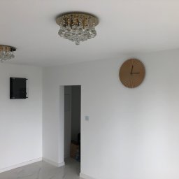 Robert Nawara - Nowoczesne wnętrze z dwoma kryształowymi lampami sufitowymi, okrągłym drewnianym zegarem na białej ścianie i marmurową podłogą.