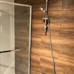 Kabina prysznicowa z chromowaną armaturą na tle ściany wyłożonej brązowymi płytkami imitującymi drewno.