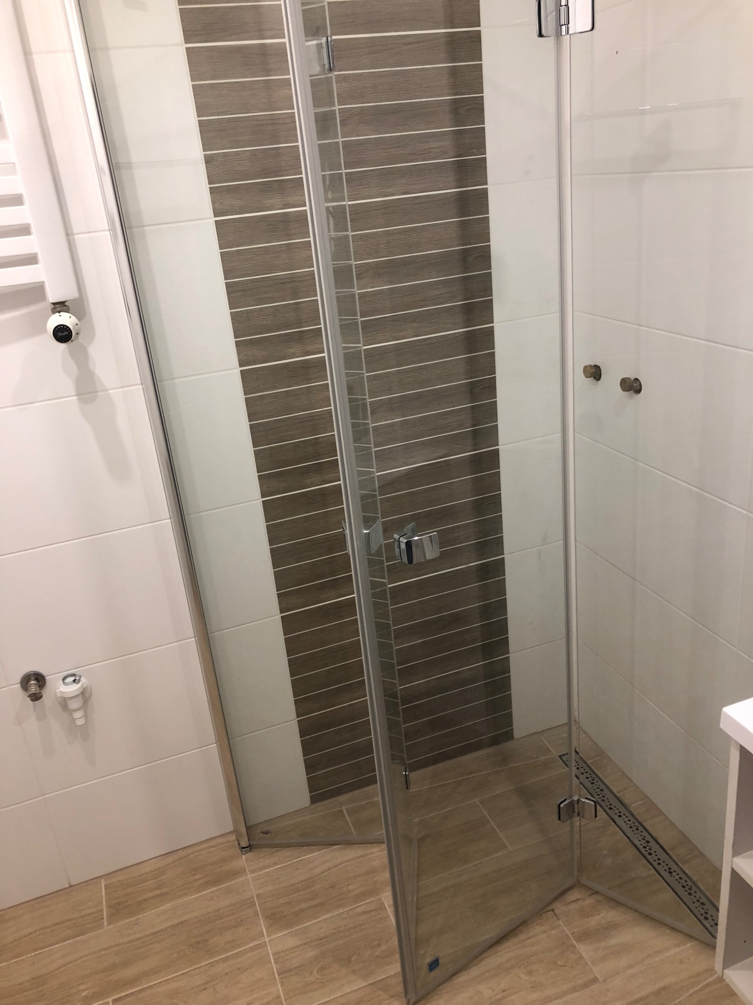 Szklana kabina prysznicowa z chromowanymi okuciami, widoczny odpływ liniowy w podłodze z płytek imitujących drewno, białe płytki na ścianach z poziomym pasem płytek w odcieniach brązu.