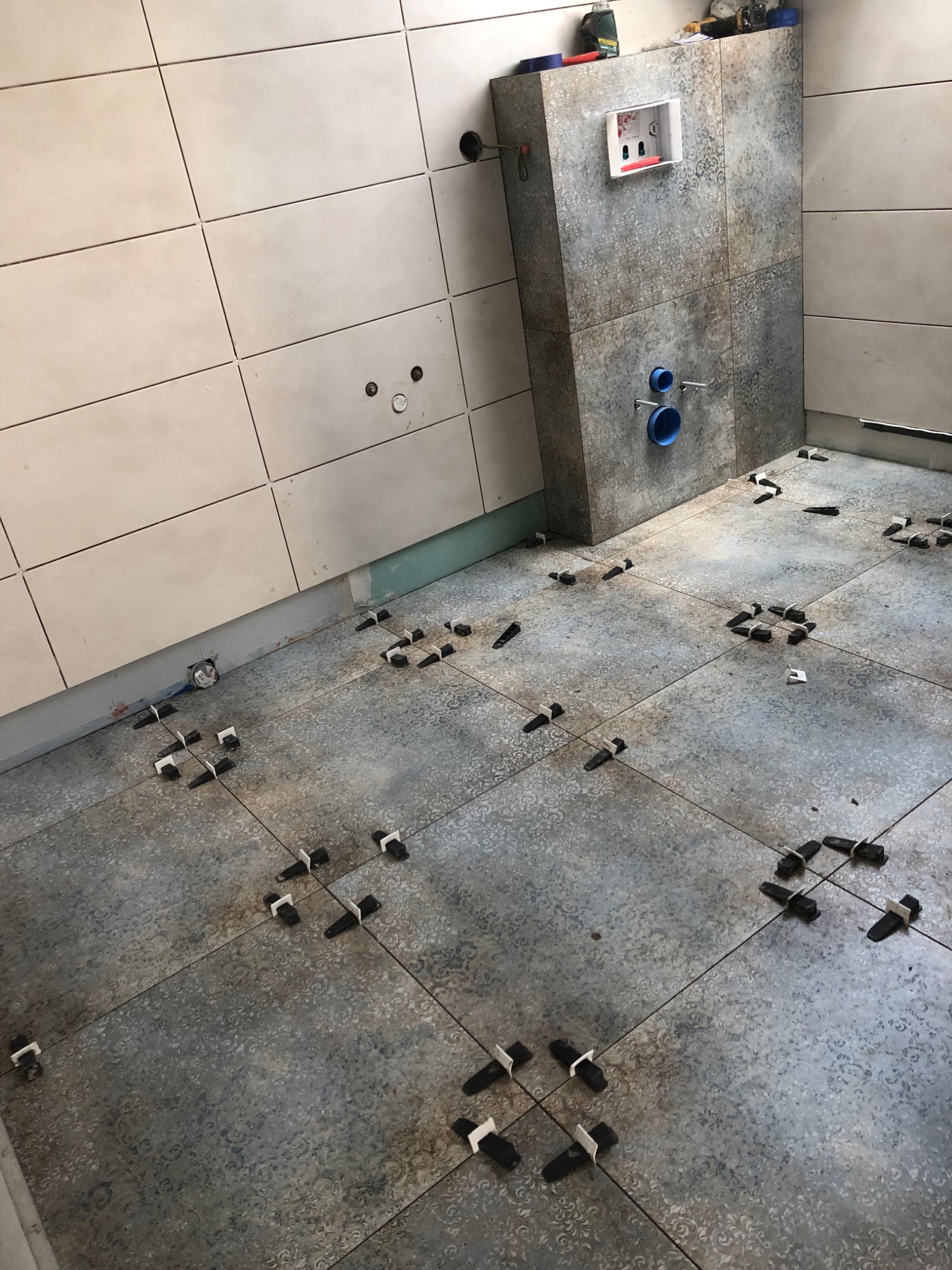 Łazienka w trakcie układania płytek podłogowych i ściennych w odcieniach beżu i szarości, z widocznymi krzyżykami dystansowymi i zabudową podtynkową WC.