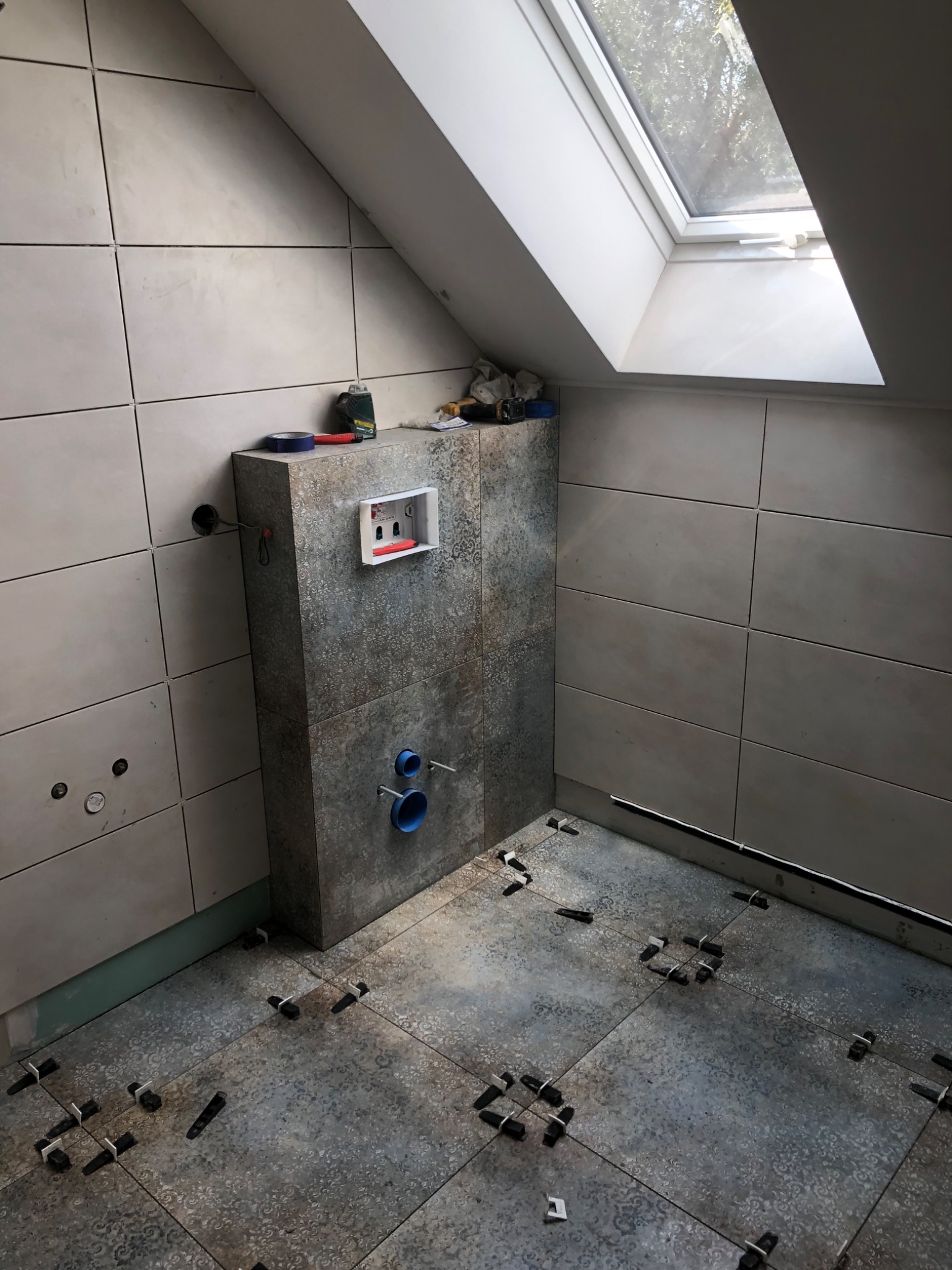 Łazienka na poddaszu w trakcie wykańczania, widoczne płytki na ścianach i podłodze z użyciem klipsów poziomujących, częściowo zabudowany stelaż podtynkowy WC, okno dachowe.