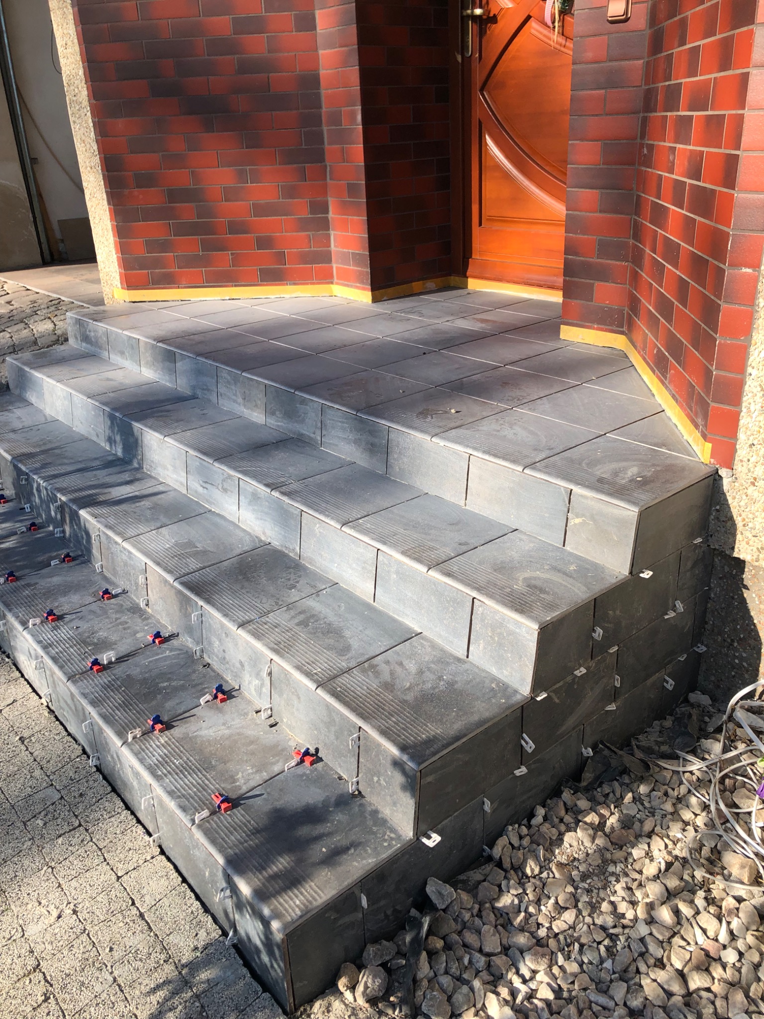 Wykończone płytkami schody wejściowe z czerwonej cegły, widoczne klipsy poziomujące, żwirowe podłoże.