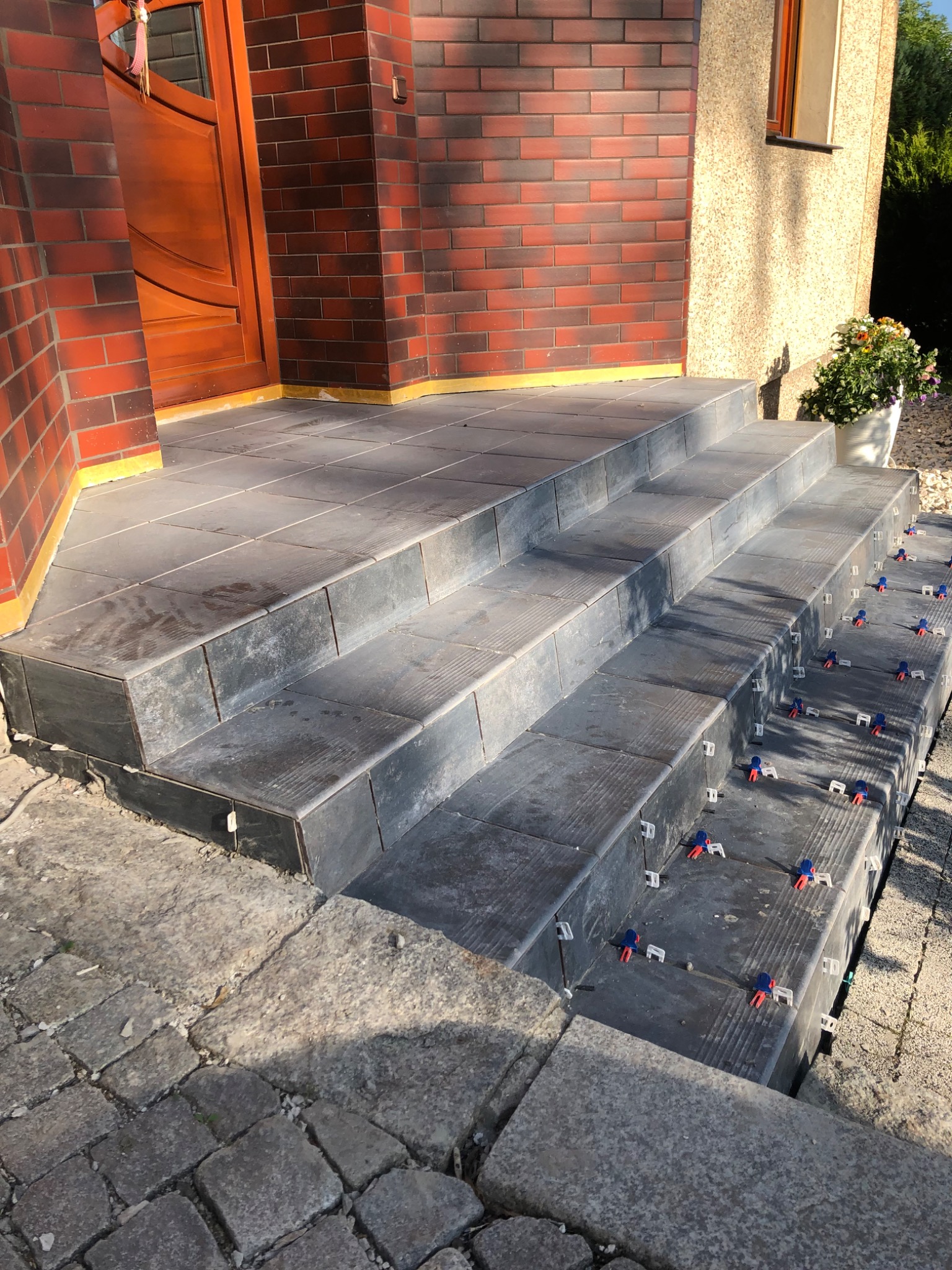 Kładzenie płytek na schodach wejściowych do domu, widoczne klipsy poziomujące, ceglana elewacja budynku w tle.