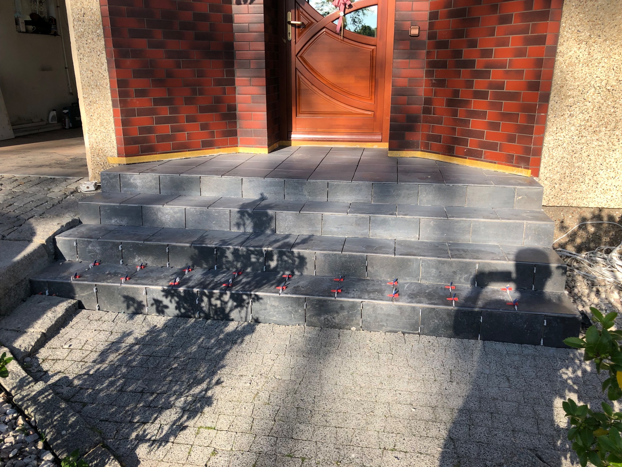 Wyłożone płytkami schody wejściowe do domu z brązową cegłą i drewnianymi drzwiami, widoczne czerwone klipsy poziomujące płytki na stopniach.