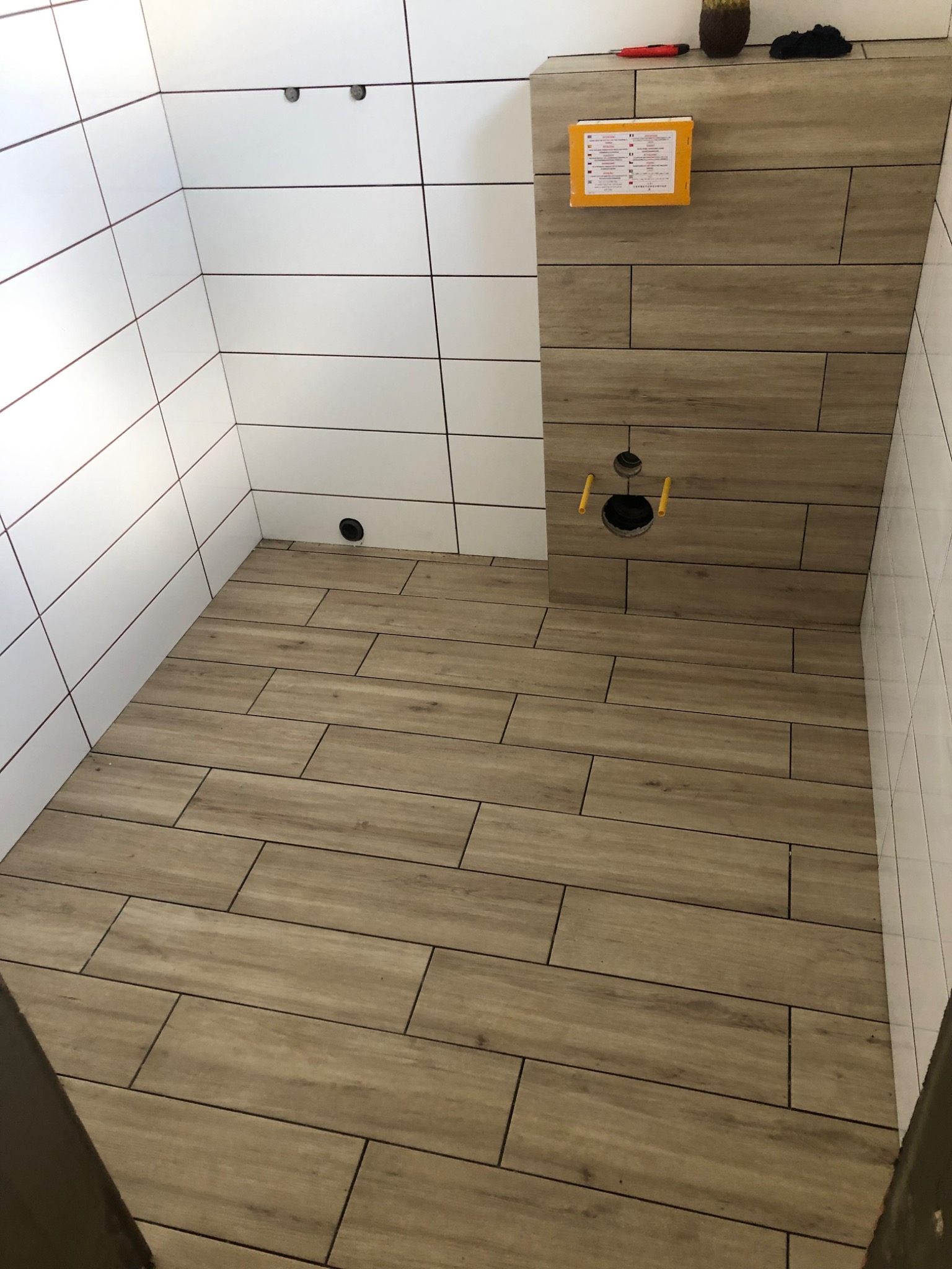 Wykończona łazienka z białymi płytkami na ścianach i drewnopodobnymi płytkami na podłodze i części ściany, widoczne otwory na armaturę, na górze panelu narzędzie i ozdobny wazon.