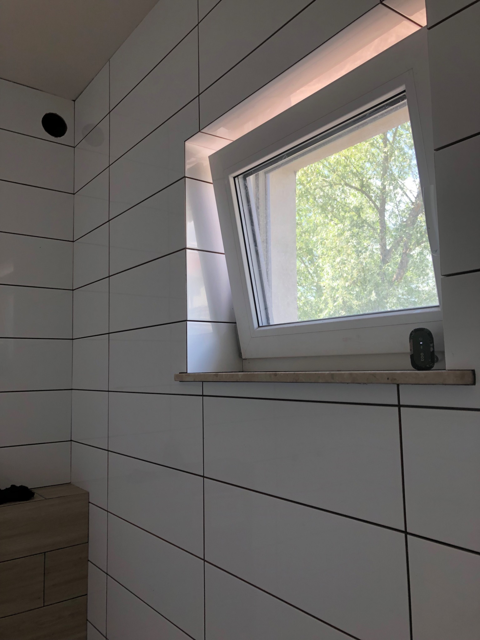 Białe, prostokątne płytki ceramiczne z ciemnymi fugami na ścianie łazienki, widoczne okno z widokiem na drzewo, mały czarny przedmiot na parapecie.