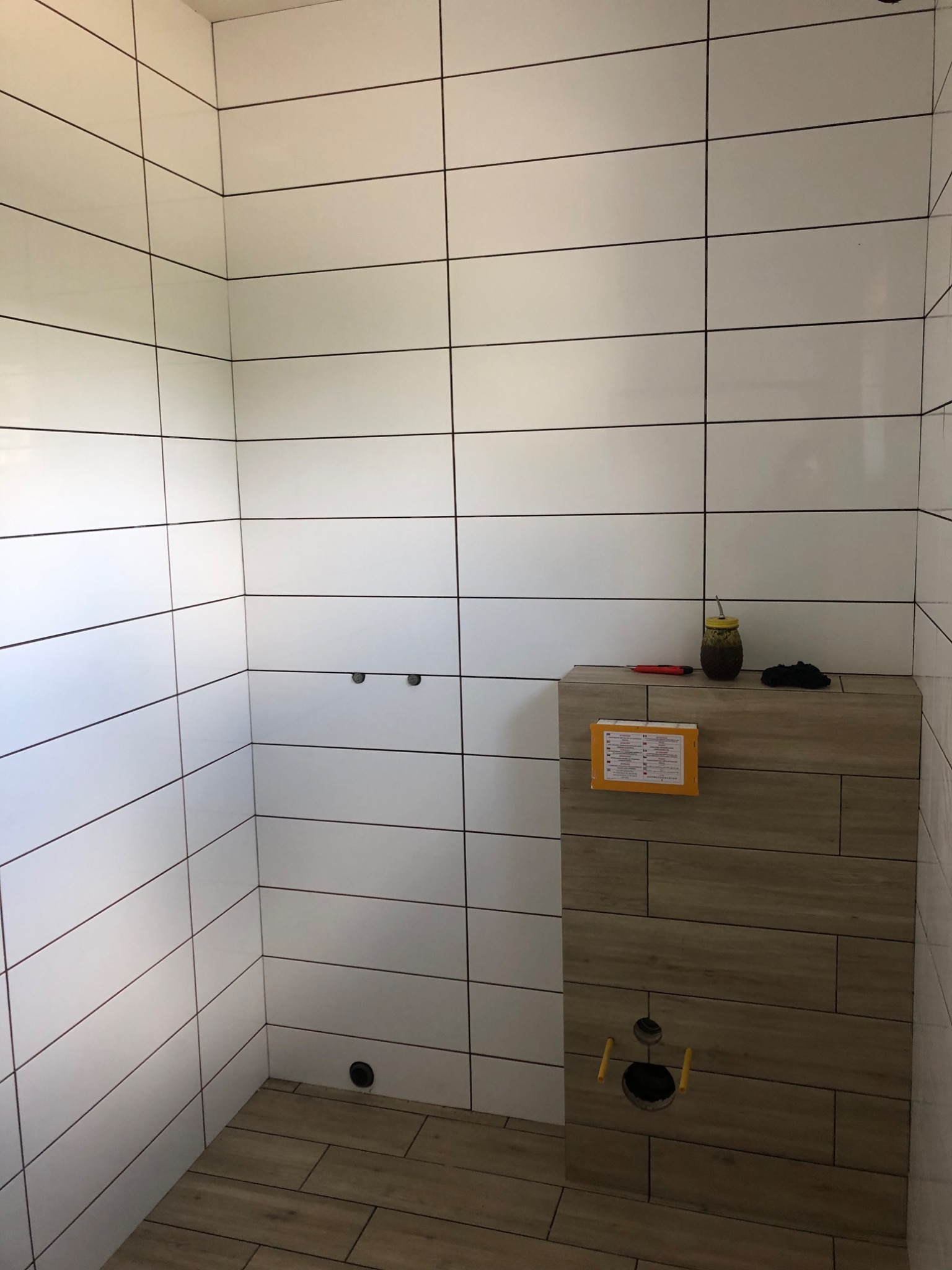 Świeżo wykafelkowana łazienka z białymi, podłużnymi płytkami i zabudową stelaża WC wykończoną drewnopodobnymi panelami; widoczny otwór na przycisk spłukujący, na zabudowie leżą narzędzia i naczynie...