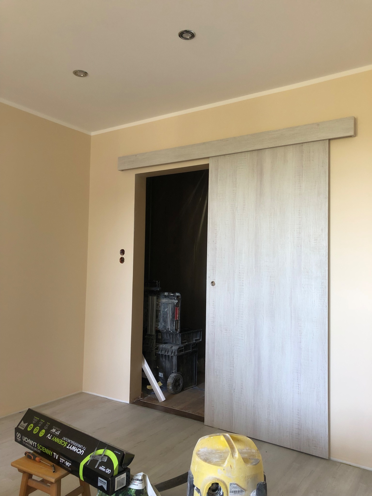 Montaż drzwi przesuwnych z szarobiałego drewna w jasnym pokoju, widoczny laser krzyżowy, skrzynki narzędziowe i opakowanie uchwytu ściennego TV na stołku.